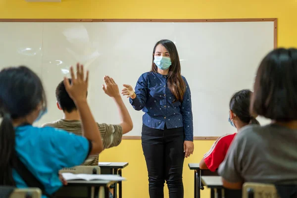 Asian teacher with protective face mask teaching her students on a class at elementary school after covid-19 quarantine and lockdown.