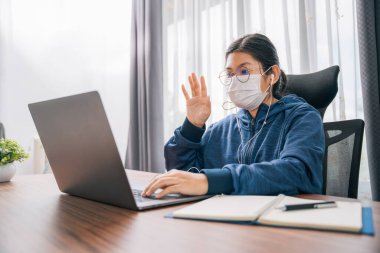 Asian young girl wear a mask gesture of greeting student learning online with headphones and laptop at home