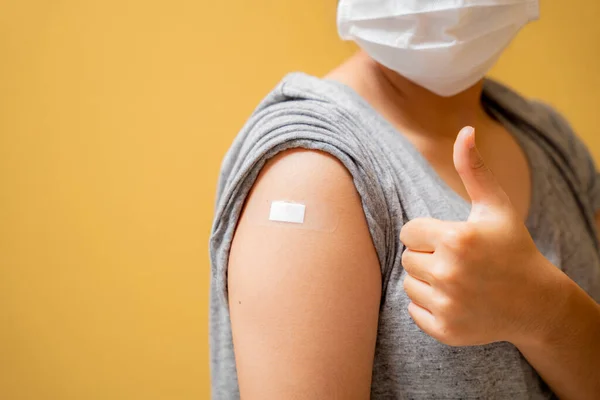 Close up of Vaccinated asian teen girl in face mask showing plaster bandage on her arm after getting covid-19 vaccine injection on yellow studio background. 