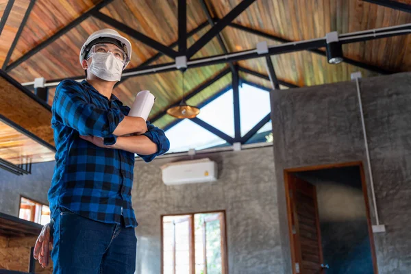 Happy asian construction engineer technician standing with arms crossed confidently after inspect the wood structure under the roof at construction site or building site of a house.