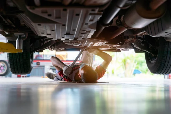 Mechanic in uniform lying down and working under car at auto service garage, car mechanic adjusting tension in vehicle suspension Element at auto repair service center, car suspension concept