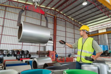 Profesyonel güvenlik donanımı işçisi, büyük, ağır çelik bir bobinin hareketini kontrol ediyor. Uzaktan kumandayla, bir fabrikada vinçle asılı duruyor..