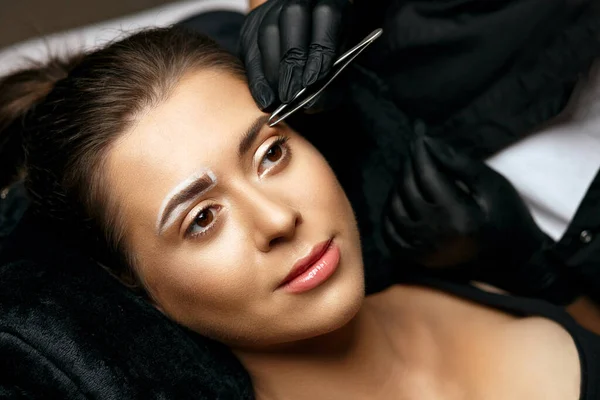 Beauty master correcting eyebrows with a tweezers before microblading procedure. Empty space