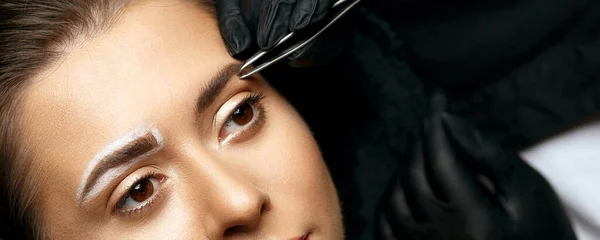 Beautician correcting eyebrows with a tweezers before permanent makeup procedure. Empty space