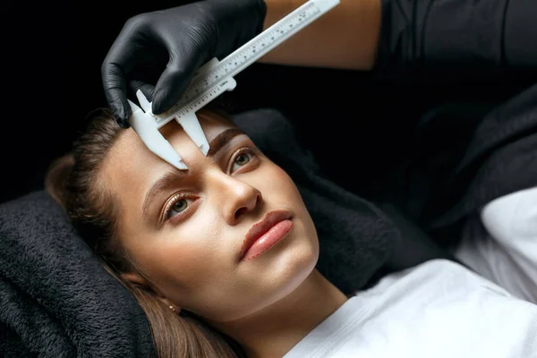 Brow master measuring eyebrows with the ruler before tattooing procedure in a beauty salon