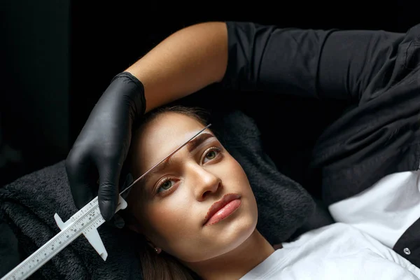 Brow stylist measuring eyebrows with the ruler before permanent makeup procedure in a beauty salon