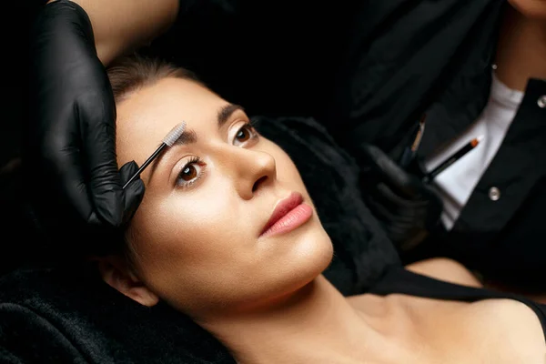 Cosmetician combing eyebrows with a brush before brow tattoo procedure. Closeup shot of a model with a lovely face