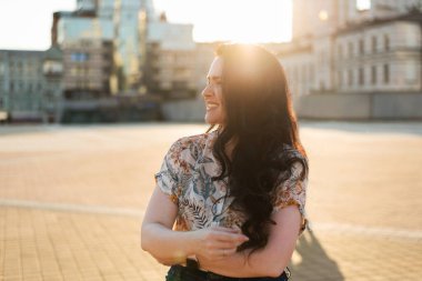 Uzun kıvırcık saçlı neşeli genç bir kadın, gömleğini giyer ve güneşli havanın tadını çıkarır.