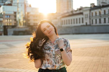 Uzun kıvırcık saçlı, gülümseyen genç kadın şehir caddesinde yürürken gömlek giyer, güneşli havanın tadını çıkarır.