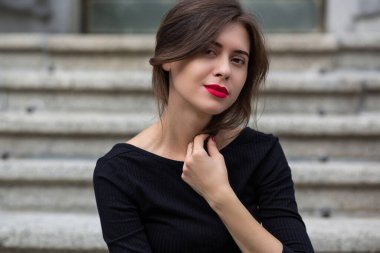 Kırmızı dudaklı esmer kadın gri beton merdivenlerde oturan siyah elbise giyiyor.