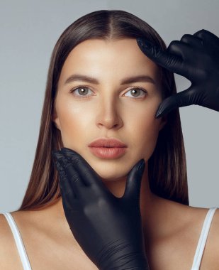 Siyah eldivenli dermatolog estetik ameliyattan önce kadının yüzünü kontrol ediyor.