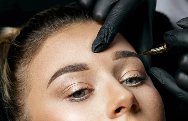 Cosmetician in gloves making brow permanent makeup to a stunning blonde woman in a beauty salon. Top view. Closeup sho