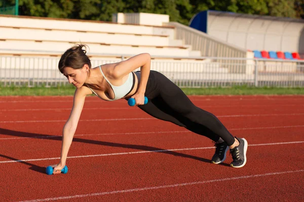Strong brunette woman in sport apparel doing exercises with dumbbells at the stadium