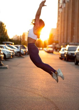 Atletik genç bir kadının altın bir akşam güneşiyle caddeye atlamasının yan görüntüsü.