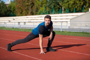 Yakışıklı sakallı adam stadyumda koşmadan önce bacak esnetiyor. Boş alan