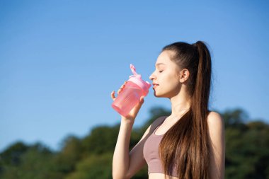 Uzun saçlı, susamış atletik kadın spor yaparken sütyenli su içiyor. Metin için boşluk