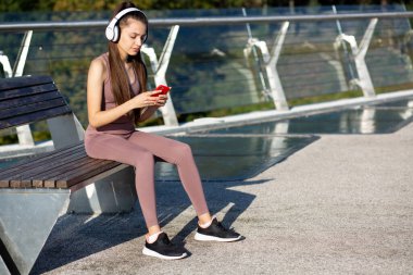 Uzun saçlı, cep telefonuna mesaj yazan ve kulaklıkla müzik dinleyen genç bir atletik kadın. Yakın çekim, spor konsepti