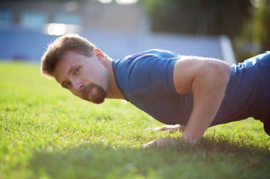 Mavi tişörtlü, çimenlikte şınav çeken kaslı bir adamın yakın plan portresi.