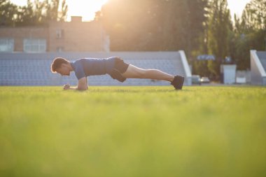 Formda bir adam spor tişörtü giyer ve pantolon giyer. Sabahları çimenlikte kalas yapar.