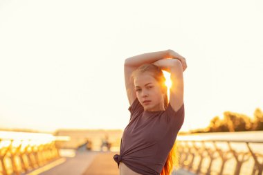 Atletik vücutlu, çekici kızıl kadın güneş doğarken köprüde poz veren spor giysiler giyiyor. Boş boşluk