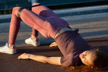 Köprüde direnç bandıyla kalça egzersizi yapan kızıl saçlı spor bir kadın. Tex için boşluk