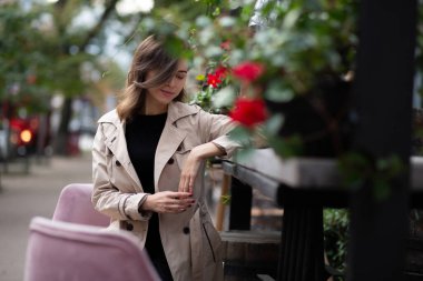Göz kamaştırıcı genç bir kadın sabahleyin sokak restoranında oturan bej bir ceket giyiyor. Dışarıda yeşil ve çiçeklerle süslenmiş. Boş boşluk
