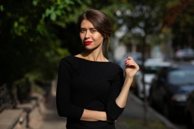 Kırmızı dudaklı zarif esmer kadın siyah elbise giyiyor. Sokakta güneş ışığıyla poz veriyor.