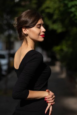 Kırmızı dudaklı, manikürlü, siyah elbiseli, caddeye güneş ışığıyla poz veren zarif genç bir kadın.