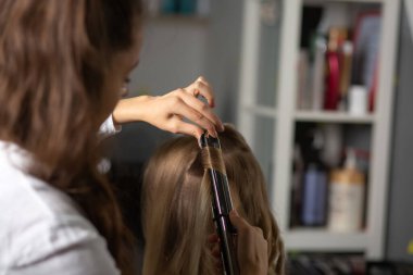 Güzellik salonunda çalışma süreci: Kadın kuaförü saç maşası olan sarışın bir kadını kıvırcık saçlı yapıyor.