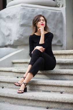 Kırmızı dudaklı ve kısa kıvırcık saçlı hoş bir genç kadının hayat tarzı merdivenlerde oturan modaya uygun siyah elbiseler giyiyor.