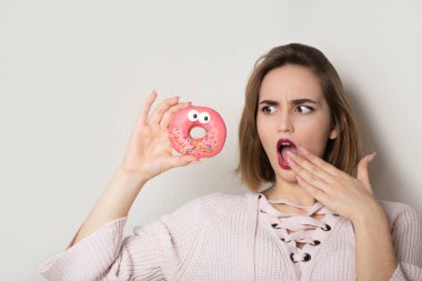 Gri arka planda pembe jöleli tatlı çörek tutan esmer kadın şaşırdı.