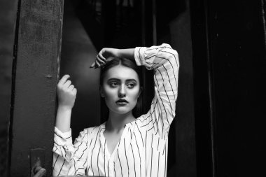 Siyah-beyaz, iç mekan portresi. Üzgün, çarpıcı genç bir kadın. Çizgili beyaz gömlek giyiyor.