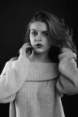 Uzun kıvırcık saçlı esmer kadın gölgede poz veren çıplak omuzlu örülmüş bir kazak giyiyor. Tek renkli çekim.