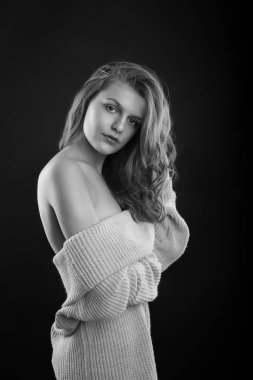 Şatafatlı esmer kadın, stüdyoda gölgede poz veren çıplak omuzlu örülü bir kazak giyiyor. Tek renkli çekim.