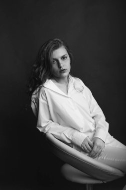 Cazibeli genç kadın beyaz gömlek ve pantolon giyip stüdyoda bir sandalyeye oturuyor. Siyah ve beyaz çekim