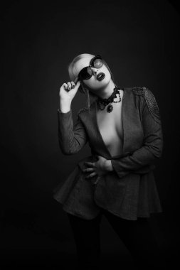 Çekici sarışın bir kadının siyah beyaz stüdyo portresi çıplak bir vücuda güneş gözlüğü ve ceket takıyor.