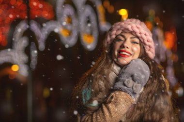 Pozitif genç kadın, kar yağarken şehirde yürürken pembe şapka, eldiven ve eşarp takıyor.