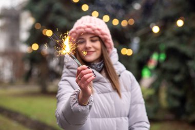 Mutlu genç kadın pembe örgü şapka ve ceket giyiyor. Noel ağacının yanındaki sokakta maytaplarla eğleniyor.
