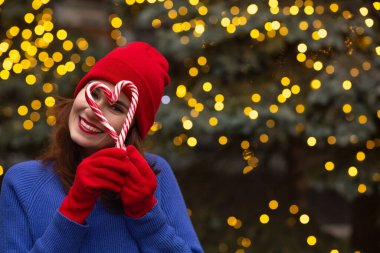 Pozitif esmer kadın kırmızı şapka ve mavi süveter giyiyor ve Noel ağacının yanında çelenklerle süslenmiş şeker tutuyor. Metin için boşluk