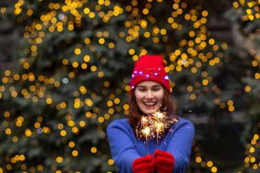Heyecanlı esmer kadın kırmızı şapka takıyor ve mavi süveter giyiyor. Noel ağacının yanında maytaplarla eğleniyor. Metin için boşluk
