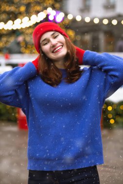 Sevimli esmer kadın Central City Meydanı 'ndaki Noel panayırında yürürken kırmızı şapka ve mavi kazak giyiyor.