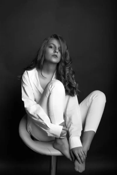 时尚的年轻女性穿着白衬衫和裤子坐在工作室的椅子上 黑白照片 — 图库照片