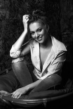 Gülümseyen esmer, doğal makyajlı kadın stüdyoda gömlek ve kot pantolon giyiyor. Siyah beyaz portre