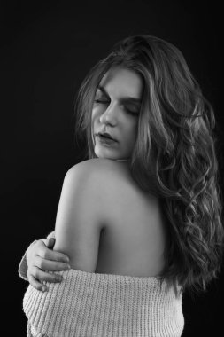 Uzun kıvırcık saçlı baştan çıkarıcı esmer kadın gölgede poz veren çıplak omuzlu örülü bir kazak giyiyor. Tek renkli çekim.