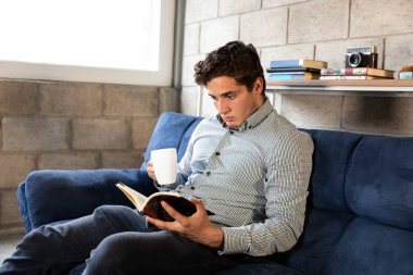 Yakışıklı ve zarif bir adam oturma odasında COVID-19 salgını sırasında kitap okuyor.