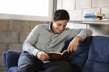 Yakışıklı ve zarif bir adam oturma odasında COVID-19 salgını sırasında kitap okuyor.