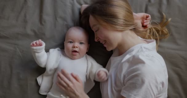 赤ちゃんを連れたお母さんが横になってカメラを上から見ている。彼女の赤ん坊と美しい若い女性。母親と娘。幸せな家族. — ストック動画