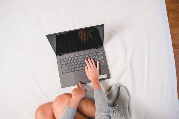 Woman\'s hands typing on laptop at home teleworking due to the virus pandemic. Girl in comfortable bed using laptop. Cenital plane