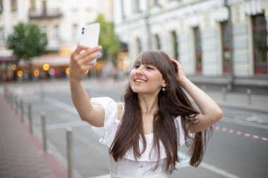 Beyaz elbiseli, uzun kahverengi saçlı, gülümseyen ve yazın arka planda cep telefonuyla selfie çeken güzel bir kızın fotoğrafı.