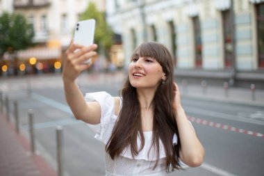 Beyaz elbiseli, uzun kahverengi saçlı güzel bir kızın yakın plan fotoğrafı. Yazın arka planda beyaz telefonun ön kamerasında gülümseyip kendi fotoğraflarını çekiyor.